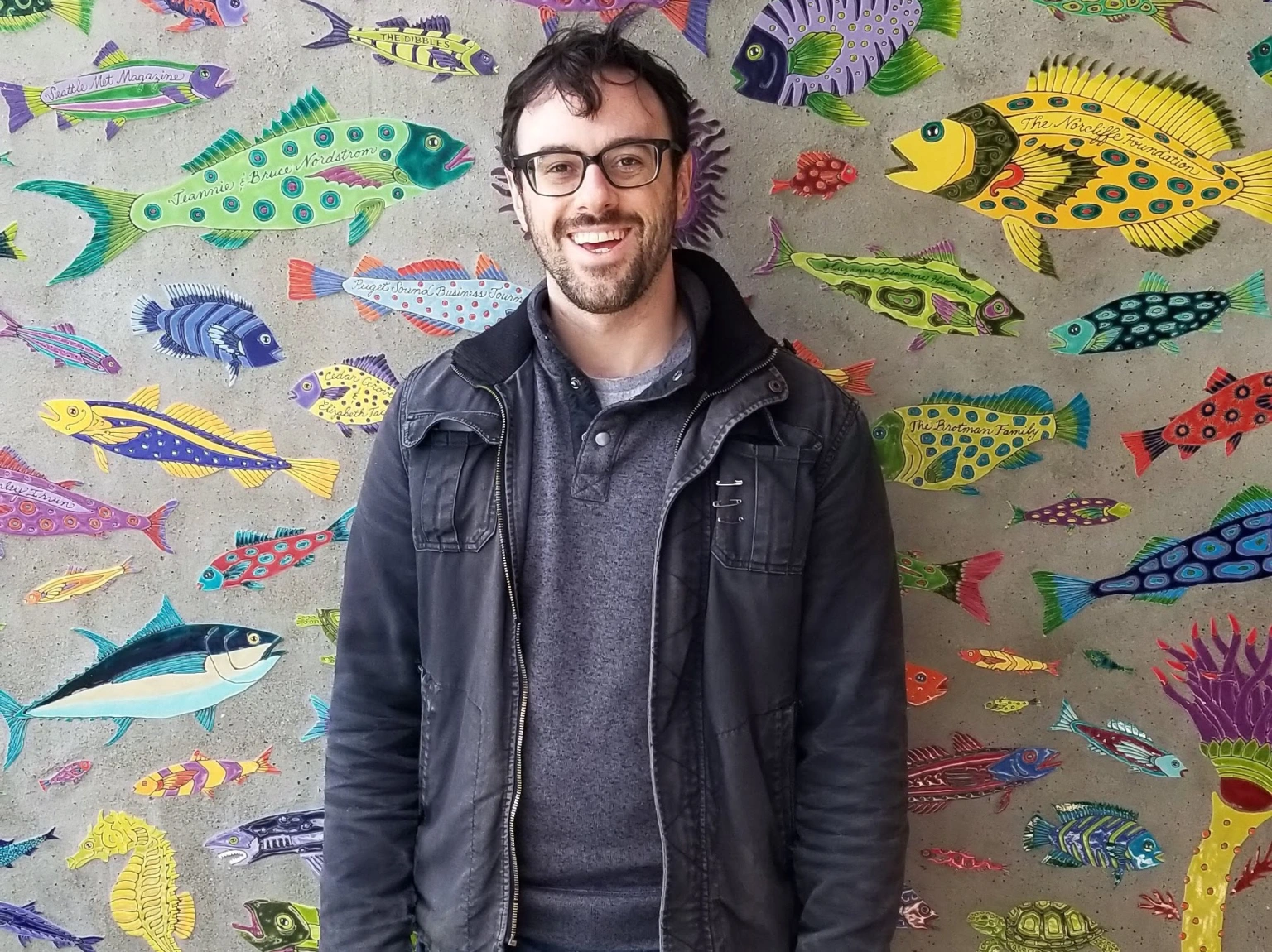 Man standing in front of color painted fish on a concrete wall. He has glasses and looks like he's mid laugh. It's very artsy vibes.